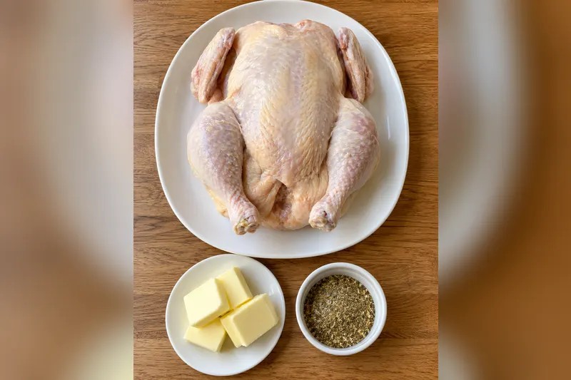 The three ingredients: a whole raw chicken on a white platter, cubes of butter and a small bowl of poultry seasoning