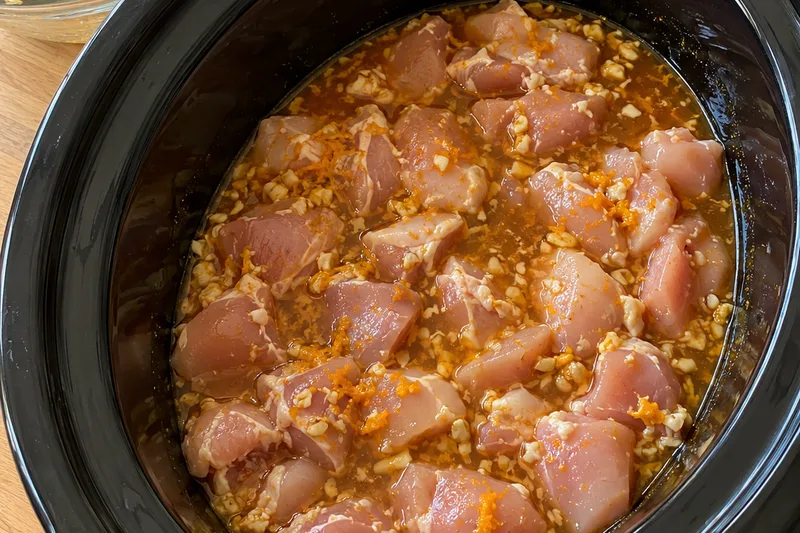 Raw bite-sized chicken pieces covered in orange sauce with garlic and zest in an oval slow cooker, before cooking