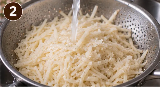 Shredded potatoes being rinsed under running water in a colander